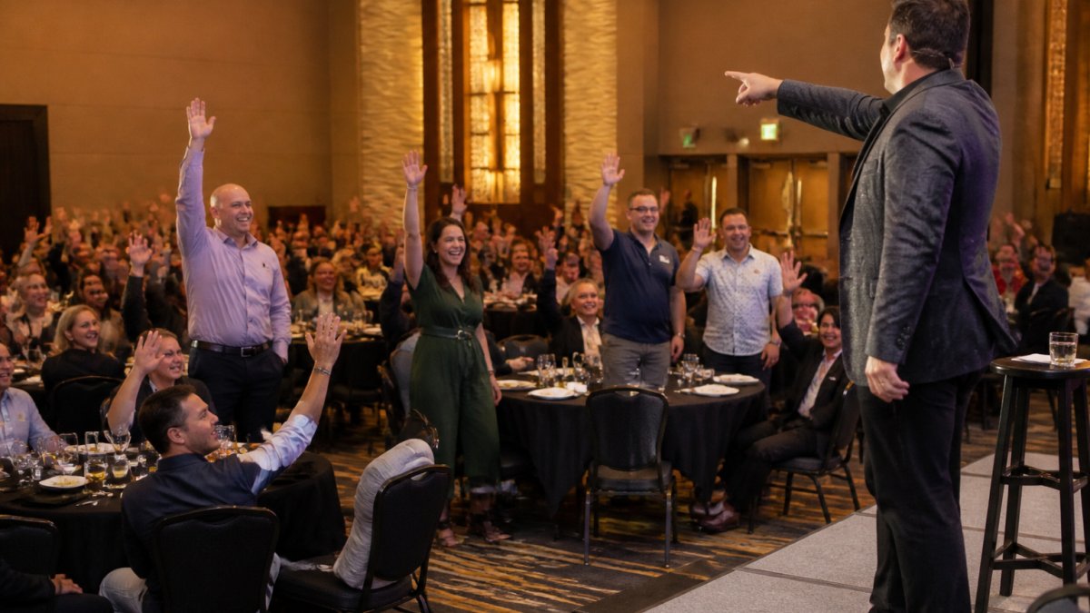 A diverse group of corporate employees laughing and raising hands as a stage performer invites volunteers up during a company event, warm stage lighting, packed banquet room with round tables