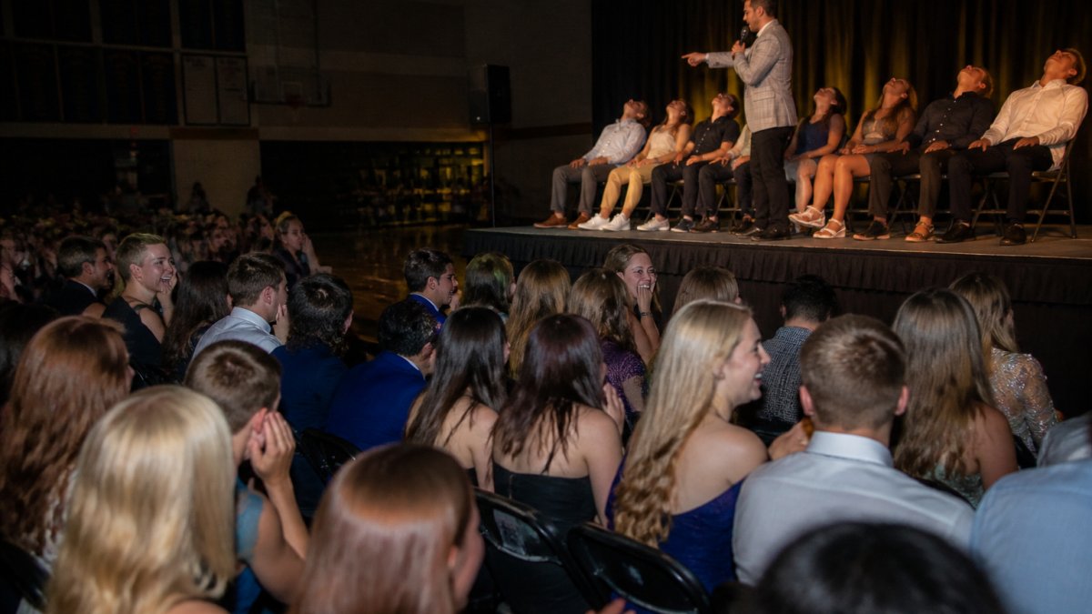 A stage hypnotist performing in front of a large audience of high school seniors at a school event, with volunteers on stage and students laughing in the crowd, warm stage lighting against a darkened gym