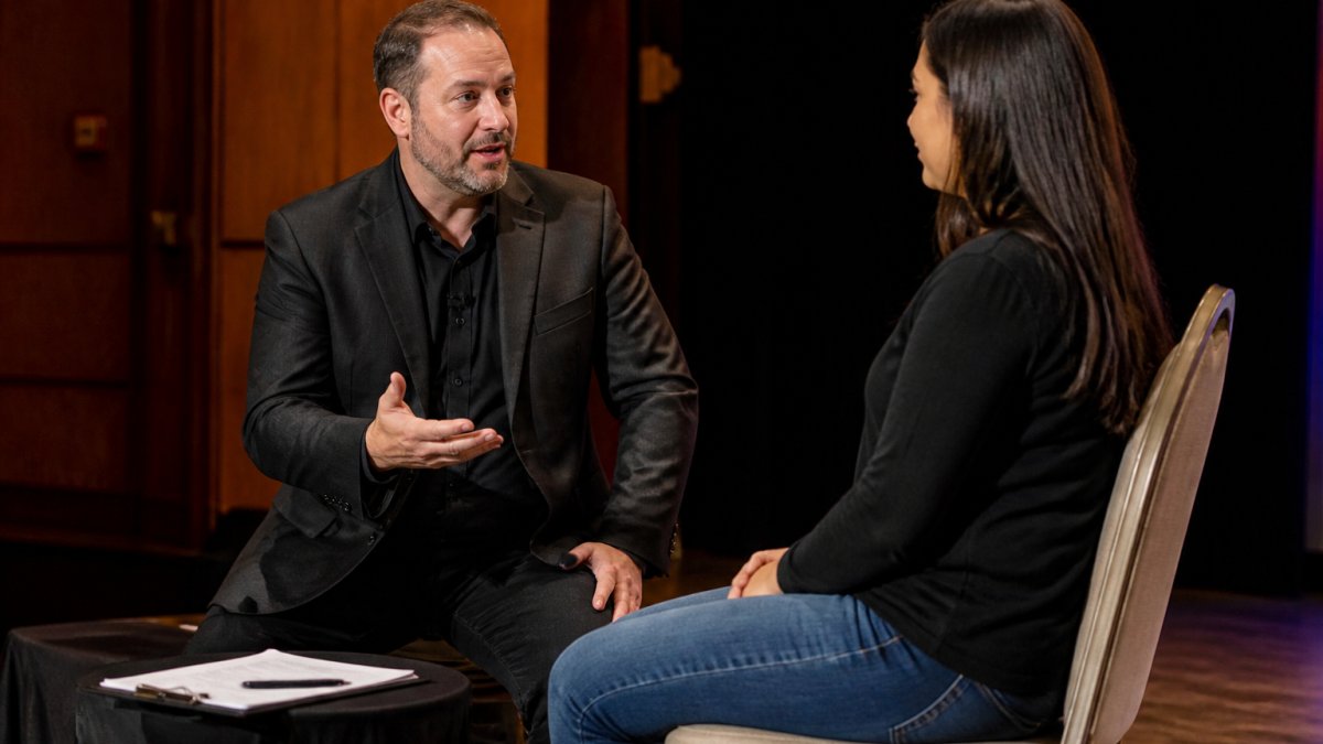 A stage hypnotist speaking calmly with a seated volunteer on a stage, a clipboard and consent forms visible on a side table, warm stage lighting