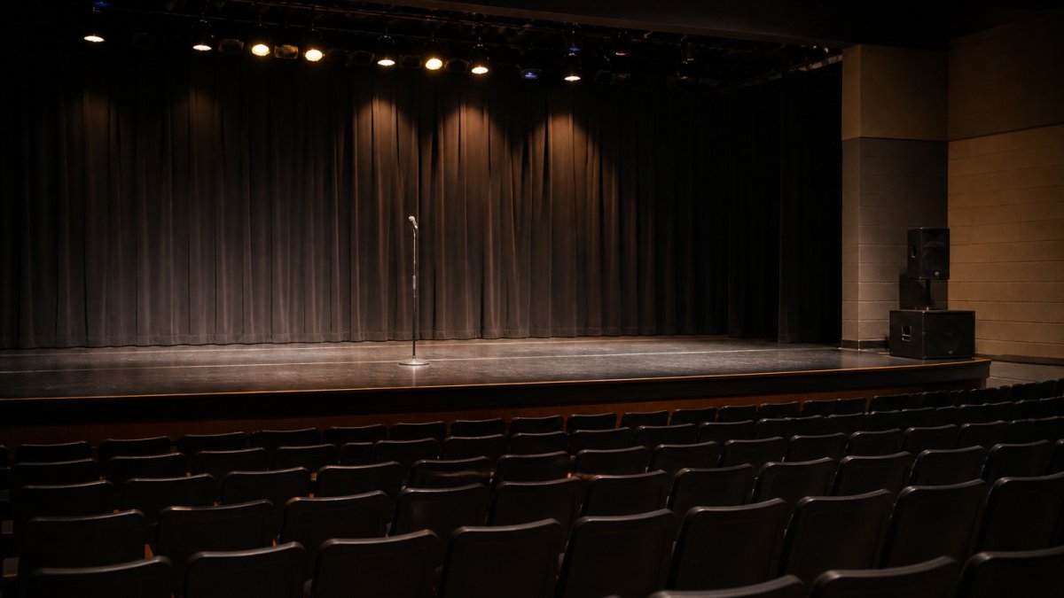 A school auditorium stage set up for a live performance, with a microphone stand, simple lighting, and rows of empty seats in the foreground